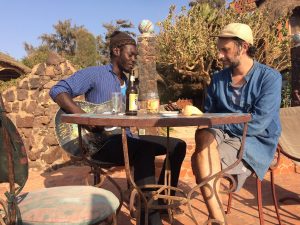 Karl-Jonas Winqvist och Babacar Diol sitter vid ett bord utomhus, Babacar spelar gitarr.