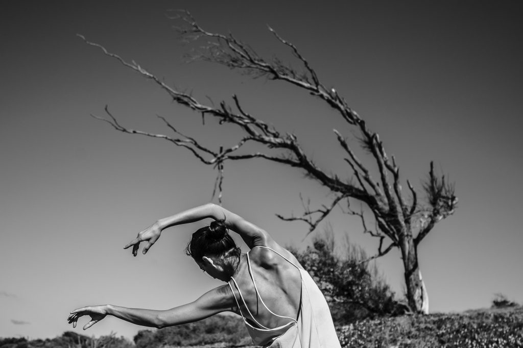 Ett svartvitt foto på en dansare som rör sig i förgrunden med armarna utsträckta uppåt åt ena hållet med ett träd i bakgrunden som lutar sig åt samma håll. 
