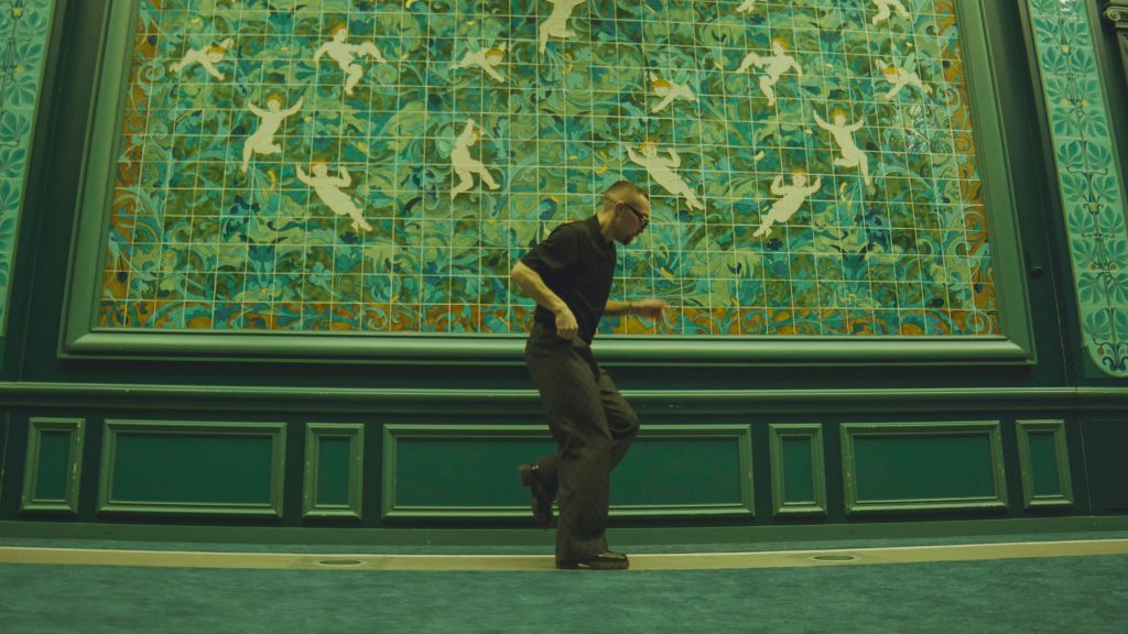 En danskonstnär i rörelse framför en stor, utsmyckad grön mosaikvägg.