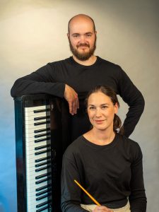 Portträttbild på två konstnärer, en sittandes och en ståendes med armen över ett lodrätt klaviatur.